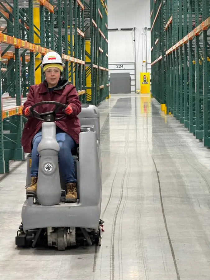 Omega crew member on a ride-on scrubber in a warehouse aisle